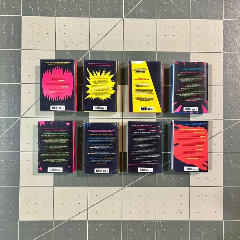 Op de afbeelding: Acht miniatuurboeken met kleurrijke ruggen en achterkanten met tekst en barcodes. De ruggen zijn donker, de achterkanten tonen ontwerpen in roze, geel en rood. De boeken zijn gerangschikt op een geruit patroon.
