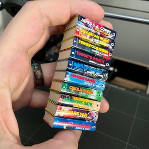 Op de afbeelding: Een stapel miniatuurboeken met kleurrijke covers. Titels zijn onder meer "Leviathan Falls" en "Persepolis Rising" van James S.A. Corey. De ruggen zijn rood, blauw, groen en oranje. De boeken worden in een hand gehouden.