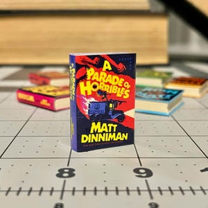 Op de afbeelding: Een miniatuurboek getiteld "A Parade of Horribles" van Matt Dinniman. De omslag heeft een rode en oranje illustratie met een blauw voertuig. Andere miniatuurboeken zijn op de achtergrond.