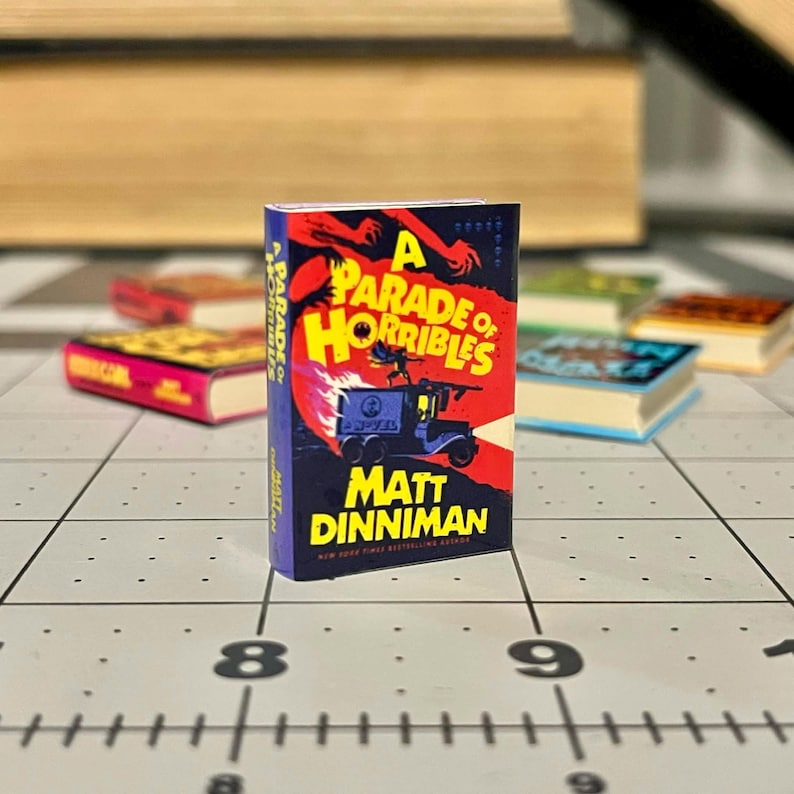 Op de afbeelding: Een miniatuurboek getiteld "A Parade of Horribles" van Matt Dinniman. De omslag heeft een rode en oranje illustratie met een blauw voertuig. Andere miniatuurboeken zijn op de achtergrond.