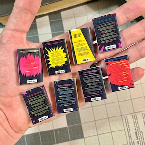 Op de afbeelding: Een verzameling miniatuurboekreplica's, elk met een uniek omslagontwerp. De ruggen en achterkanten hebben levendige kleuren zoals roze, geel en blauw, met verschillende grafische elementen en tekst. De boeken worden in een hand gehouden.