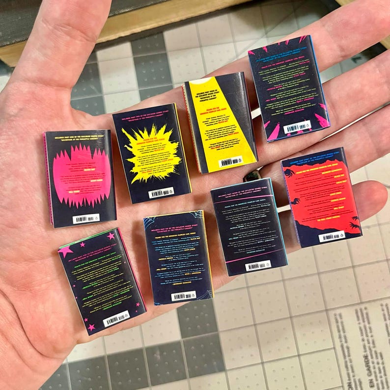 Op de afbeelding: Een verzameling miniatuurboekreplica's, elk met een uniek omslagontwerp. De ruggen en achterkanten hebben levendige kleuren zoals roze, geel en blauw, met verschillende grafische elementen en tekst. De boeken worden in een hand gehouden.