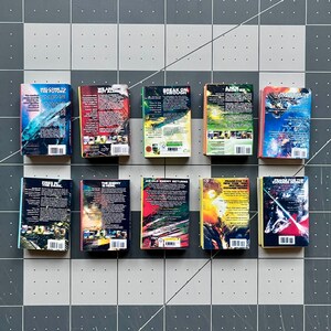 Op de afbeelding: Een verzameling van tien miniatuurboeken met kleurrijke covers en titels als "Welcome to the Future" en "Break On Through." De boeken zijn gerangschikt op een grijs en wit geblokt oppervlak, waarbij hun levendige ontwerpen en tekst worden getoond.