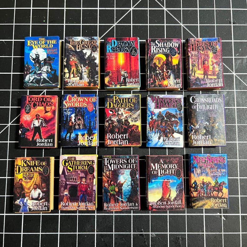 May include: A collection of 14 miniature hardcover books with colorful covers featuring fantasy illustrations and titles like "The Eye of the World", "The Great Hunt", "Dragon Reborn", "The Shadow Rising", "The Fires of Heaven", "Lord of Chaos", "Crown of Swords", "The Path of Daggers", "Winter's Heart", "Crossroads of Twilight", "Knife of Dreams", "The Gathering Storm", "Towers of Midnight", "A Memory of Light", and "New Spring".