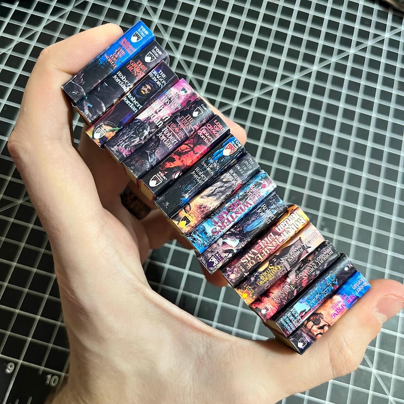 May include: A stack of 14 miniature paperback books with fantasy covers featuring dragons, wolves, and other mythical creatures. The books are arranged vertically with the spines facing out. The titles include "The Dragon Reborn", "The Great Hunt", "The Fires of Heaven", "Lord of Chaos", "A Crown of Swords", "The Path of Daggers", "Winter's Heart", "Crossroads of Twilight", "Knife of Dreams", "The Gathering Storm", "Towers of Midnight", "A Memory of Light", "The Shadow Rising", and "The Eye of the World".