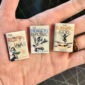 May include: Three miniature book replicas. The titles are "The Poppy War", "The Dragon Republic", and "The Burning God". Each book has a white background with text and illustrations. The books are held in a hand.