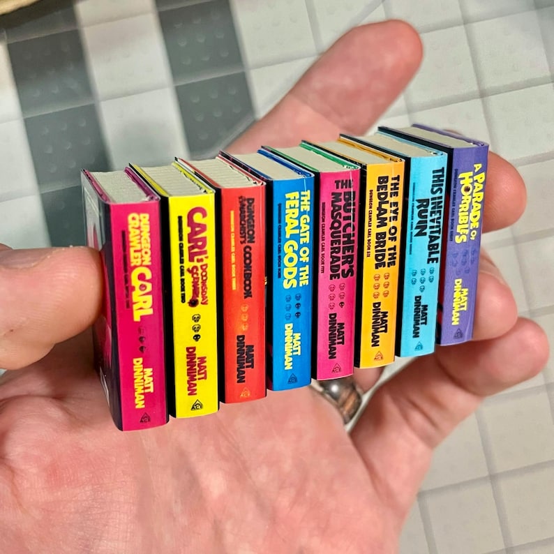 Op de afbeelding: Een verzameling miniatuurboeken met kleurrijke ruggen. Titels zijn onder meer "Dungeon Crawler Carl" en "A Parade of Horribles". Elk boek heeft een unieke kleur en titel, met de naam van de auteur onderaan.
