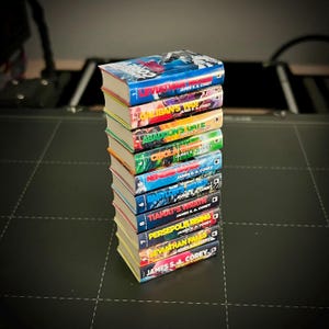 Op de afbeelding: Een stapel van tien boeken met kleurrijke covers, met titels als "Leviathan Falls" en "Tiamat's Wrath" van James S.A. Corey. De boeken hebben vergeelde pagina's en zijn op een donkere ondergrond gestapeld. De titels staan op de ruggen.