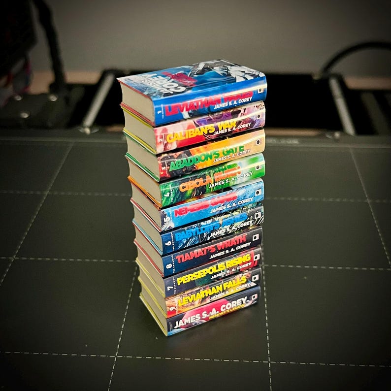 Op de afbeelding: Een stapel van tien boeken met kleurrijke covers, met titels als "Leviathan Falls" en "Tiamat's Wrath" van James S.A. Corey. De boeken hebben vergeelde pagina's en zijn op een donkere ondergrond gestapeld. De titels staan op de ruggen.