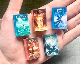 May include: Five miniature books from the Percy Jackson & The Olympians series, held in a hand. The books have colourful covers with titles including "The Lightning Thief" and "The Sea of Monsters". The books are small and detailed.
