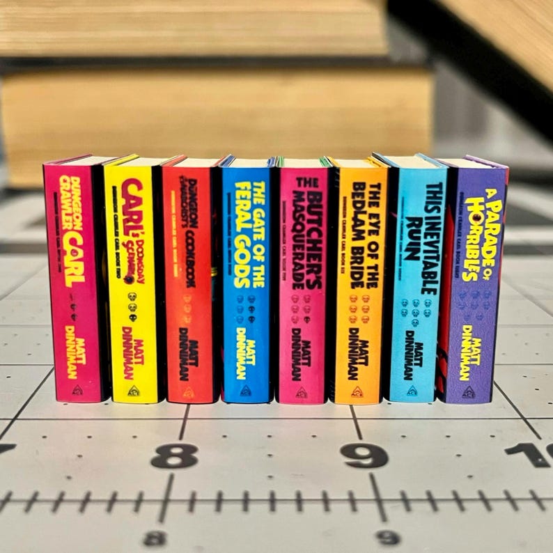 Op de afbeelding: Een verzameling miniatuurboeken met kleurrijke ruggen. Titels zijn onder meer "Dungeon Crawler Carl", "Carl", "Dungeon Cookbook", "The Gate of the Feral Gods", "The Butcher's Masquerade", "The Bedlam Bride", "This Inevitable Ruin" en "A Parade of Horribles".
