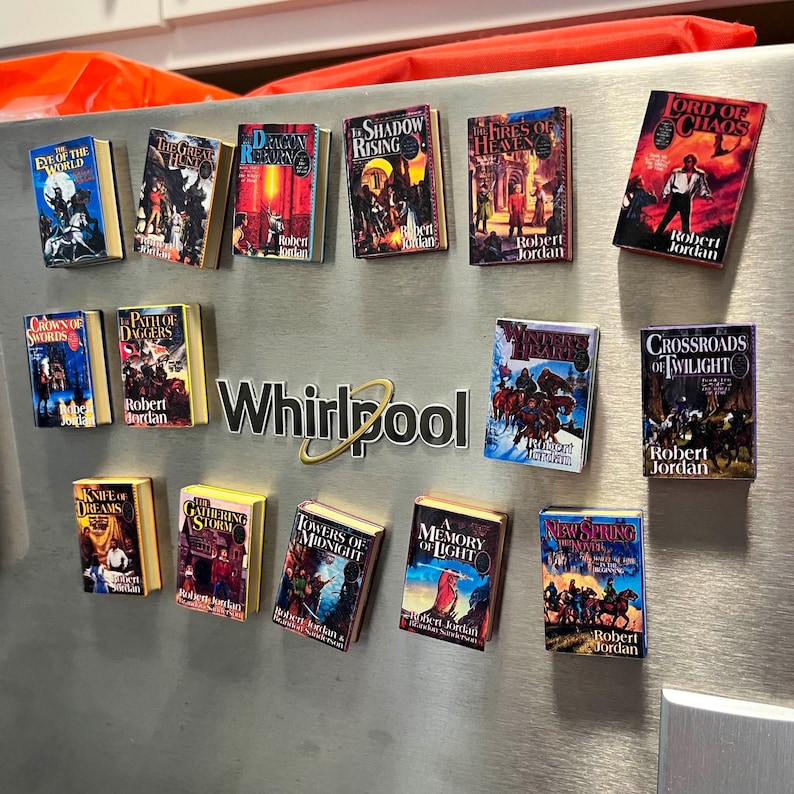 May include: A collection of 14 miniature book covers featuring the Wheel of Time series by Robert Jordan. The covers are arranged in a grid on a silver refrigerator. The covers feature colorful illustrations and text, including titles like "Eye of the World", "The Shadow Rising", and "The Fires of Heaven".
