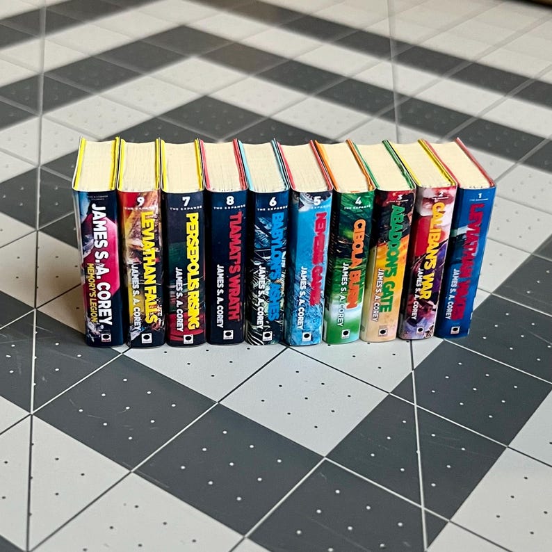 Op de afbeelding: Een rij miniatuurboeken met kleurrijke ruggen, waaronder titels als "Leviathan Falls" en "Persepolis Rising" van James S.A. Corey. De boeken hebben gele bovenkanten en gevarieerde covers.
