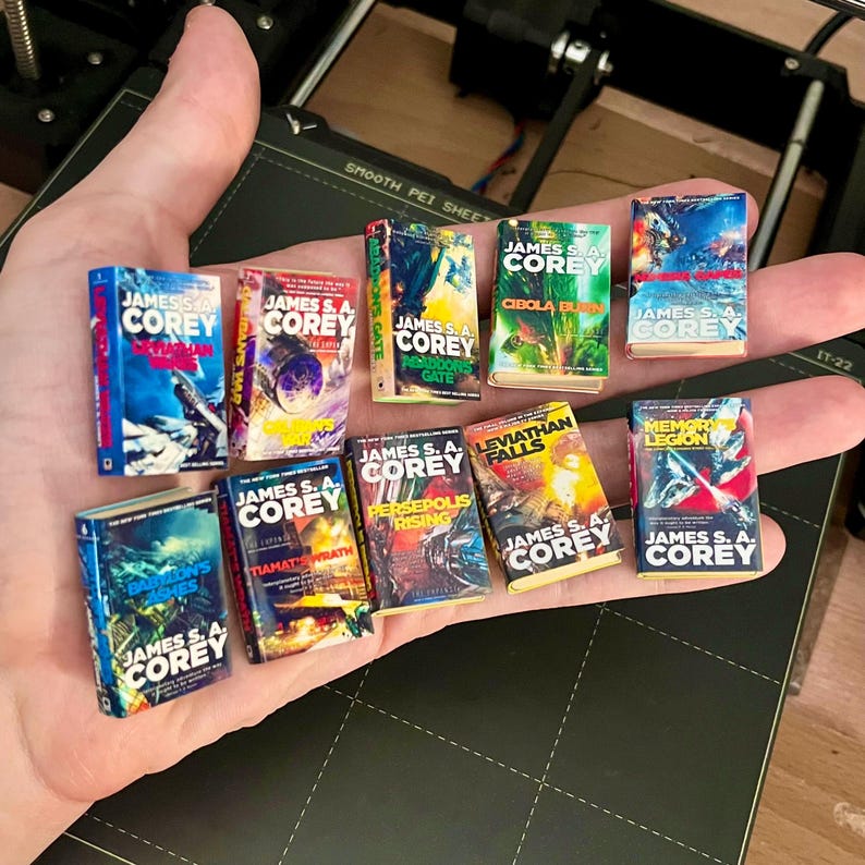 Op de afbeelding: Een verzameling miniatuurboeken, elk met de naam van de auteur "JAMES S. A. COREY" en verschillende titels. De boekcovers tonen levendige kleuren en sciencefiction-kunstwerken. De boeken worden in een hand gehouden.