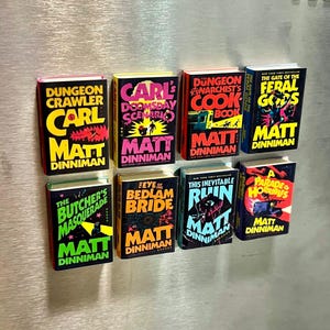 Op de afbeelding: Een verzameling miniatuurboekreplica's met kleurrijke covers. Titels zijn onder meer "Dungeon Crawler", "Carl's Doomsday Scenario" en "The Feral Gods". Elk boek toont de naam van de auteur, Matt Dinniman.