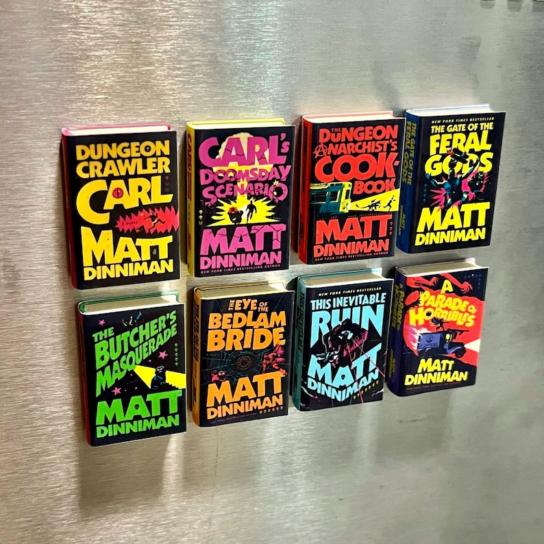 Op de afbeelding: Een verzameling miniatuurboekreplica's met kleurrijke covers. Titels zijn onder meer "Dungeon Crawler", "Carl's Doomsday Scenario" en "The Feral Gods". Elk boek toont de naam van de auteur, Matt Dinniman.