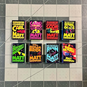 Op de afbeelding: Acht miniatuurboeken met kleurrijke covers zijn gerangschikt op een raster. De boeken hebben titels als "Dungeon Crawler" en "The Butcher's Masquerade", met de naam van de auteur, Matt Dinniman. De boeken liggen op een grijs en wit geruit oppervlak.