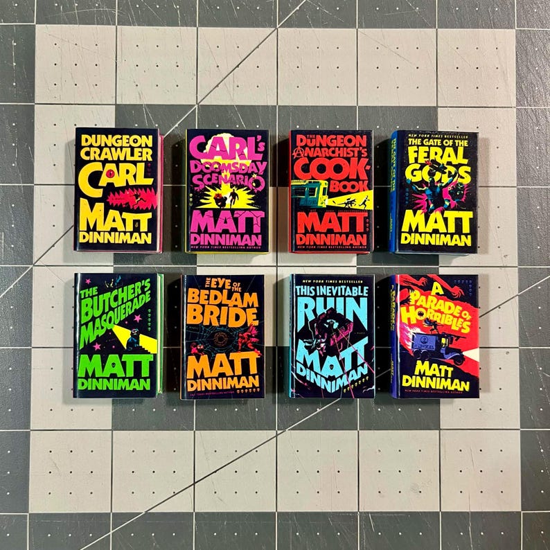 Op de afbeelding: Acht miniatuurboeken met kleurrijke covers zijn gerangschikt op een raster. De boeken hebben titels als "Dungeon Crawler" en "The Butcher's Masquerade", met de naam van de auteur, Matt Dinniman. De boeken liggen op een grijs en wit geruit oppervlak.