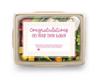 Può includere: Un contenitore di plastica trasparente pieno di verdure fresche, tra cui broccoli, carote, piselli e funghi. Un biglietto rosa e bianco con il testo "Congratulazioni per il tuo nuovo bambino! Fatto apposta per te, così avrai meno tempo in cucina e più tempo per coccole con il bambino!" è posto sopra le verdure.
