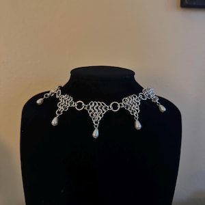 Puede incluir: Un collar de malla de plata con un colgante en forma de lágrima. El collar tiene un diseño delicado y está adornado con cinco gotas de perlas.
