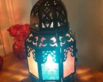May include: A black metal lantern with colorful stained glass panels. The lantern has a decorative design and is lit from within, casting a warm glow.