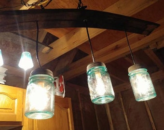 May include: A rustic wooden beam chandelier with four glass mason jars as light fixtures. The jars are clear glass with a light blue tint and have a metal lid. The jars are hanging from black metal chains.