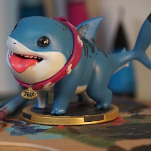 May include: A blue and white shark figurine with a pink collar and a gold tag that says "Jeff". The figurine is standing on a gold base and is on a wooden surface.