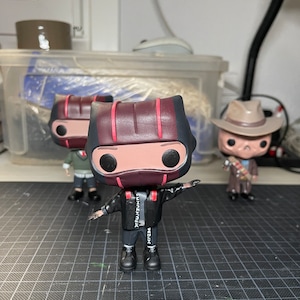 May include: Three black and red pop vinyl figures. The centre figure is wearing a black jacket with white text that says "BEZUX" and "EXXZEMAUX". The figure is also wearing black trousers and black shoes. The figure has a red and black striped helmet with a red visor. The figure has black eyes and a white face.