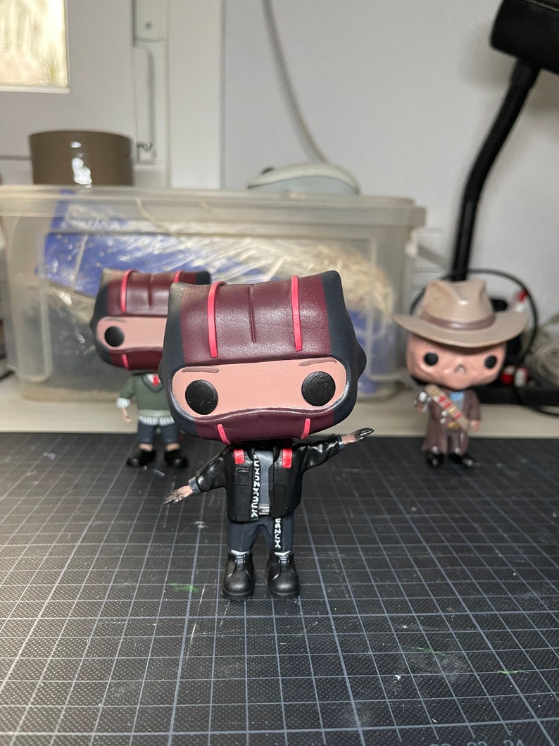 May include: Three black and red pop vinyl figures. The centre figure is wearing a black jacket with white text that says "BEZUX" and "EXXZEMAUX". The figure is also wearing black trousers and black shoes. The figure has a red and black striped helmet with a red visor. The figure has black eyes and a white face.