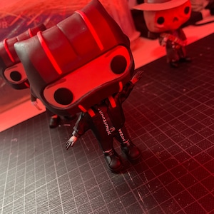 May include: Black and red vinyl figure with a black and white outfit and red stripes on the head. The figure is standing on a black grid surface. The figure has a white inscription on the back that reads "VRZUX", "VRZUX", and "YUMMZUX".