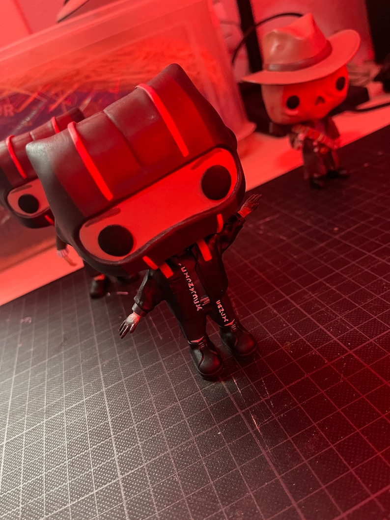 May include: Black and red vinyl figure with a black and white outfit and red stripes on the head. The figure is standing on a black grid surface. The figure has a white inscription on the back that reads "VRZUX", "VRZUX", and "YUMMZUX".