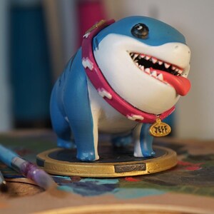 May include: A blue and white shark figurine wearing a pink collar with a gold tag that reads "Jeff". The figurine is standing on a wooden surface.