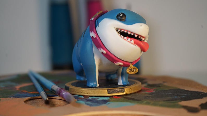 May include: A blue and white shark figurine wearing a pink collar with a gold tag that reads "Jeff". The figurine is standing on a wooden surface.