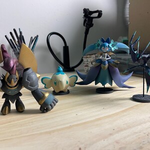 May include: Four painted figurines on a wooden surface. A black and gold robot, a white and blue elephant, a blue and purple fairy, and a black and green creature.