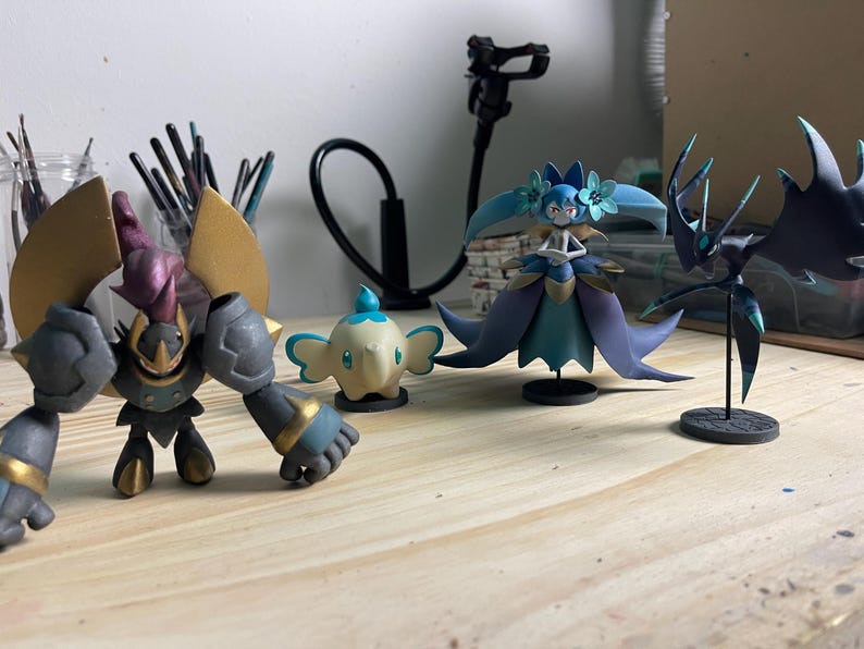 May include: Four painted figurines on a wooden surface. A black and gold robot, a white and blue elephant, a blue and purple fairy, and a black and green creature.