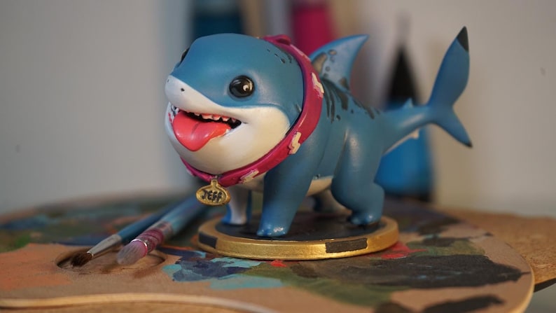May include: A blue and white shark figurine with a pink collar and a gold tag that says "Jeff". The figurine is standing on a gold base and is on a wooden surface.