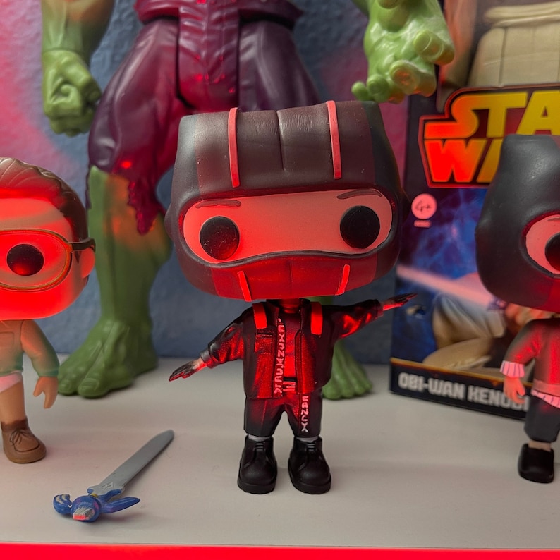 May include: A red and black pop vinyl figure of a person wearing a black jacket with red accents and a black helmet with red stripes. The figure is standing on a white surface.