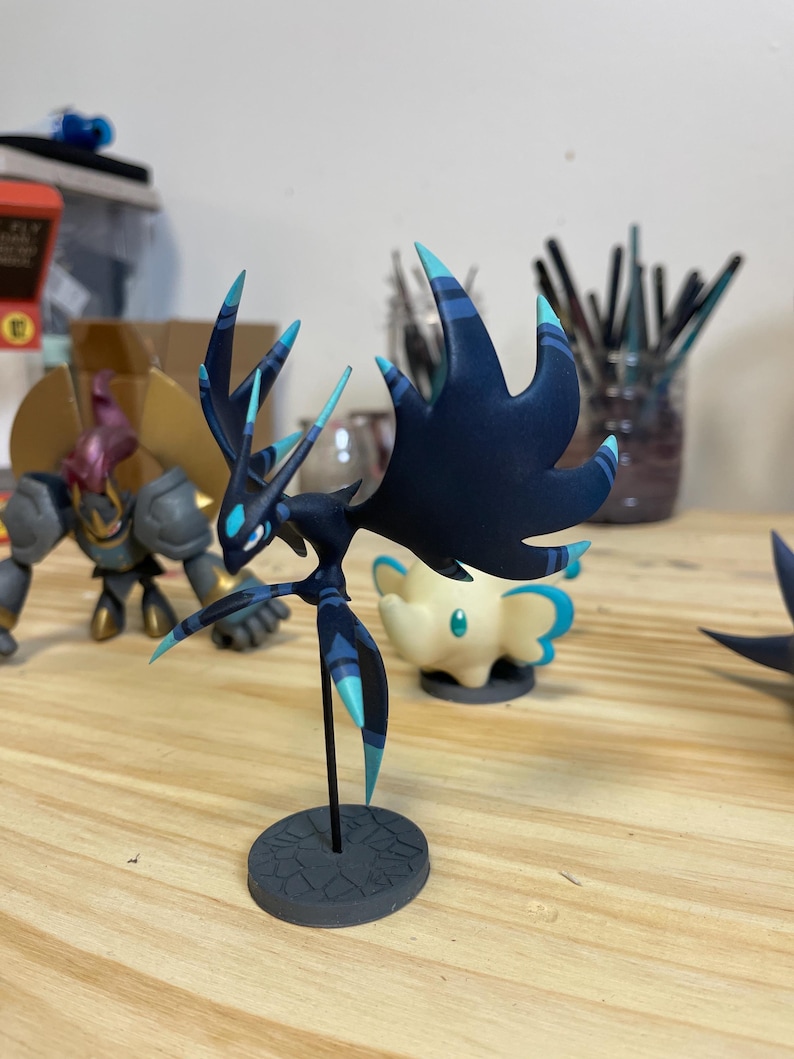 May include: A dark blue and teal colored figurine of a mythical creature with large wings and sharp points. The figurine is standing on a gray base.