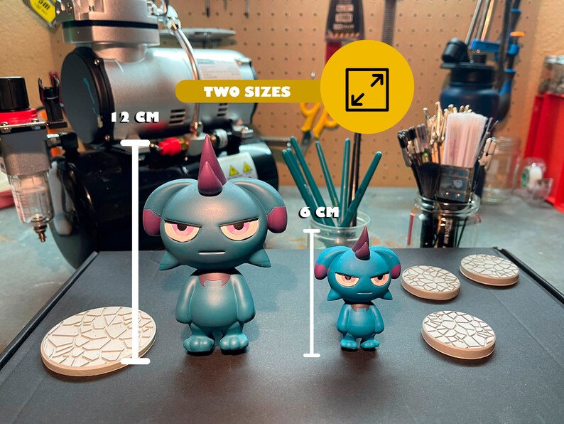 May include: Two teal blue cartoon figures with purple horns and angry expressions. The figures are standing on white bases with a textured design. The larger figure is 12 cm tall and the smaller figure is 6 cm tall. "TWO SIZES" is written in yellow on a black background.