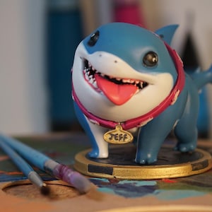 May include: A blue and white shark figurine with a pink collar and a name tag that says "Jeff". The figurine is on a gold base and is sitting on a wooden surface.
