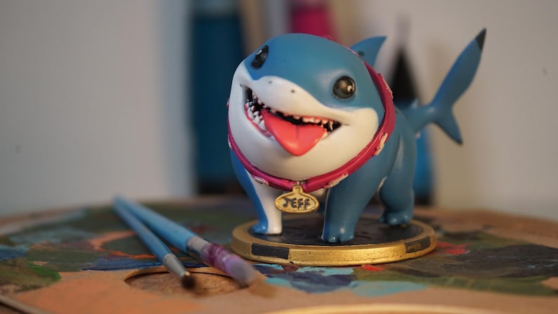 May include: A blue and white shark figurine with a pink collar and a name tag that says "Jeff". The figurine is on a gold base and is sitting on a wooden surface.