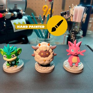 May include: Three hand-painted figurines of cartoon characters. The characters are a green monkey-like creature, a white sheep-like creature, and a pink cat-like creature. The figurines are on a gray surface. "HAND PAINTED" is written on a yellow background with a paintbrush icon.