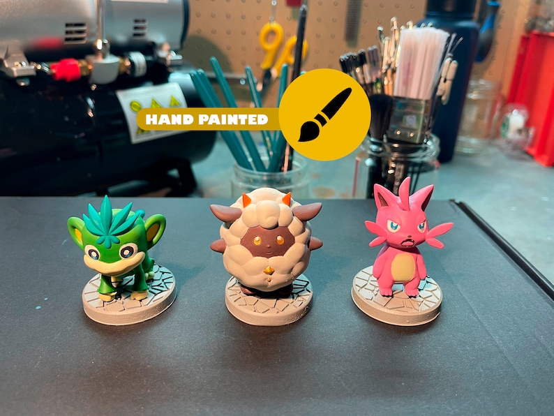 May include: Three hand-painted figurines of cartoon characters. The characters are a green monkey-like creature, a white sheep-like creature, and a pink cat-like creature. The figurines are on a gray surface. "HAND PAINTED" is written on a yellow background with a paintbrush icon.