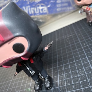 May include: A black and red vinyl figure wearing a black hooded jacket and black trousers. The figure has a black and white graphic on the jacket that says "Unbreakable". The figure is wearing black shoes.
