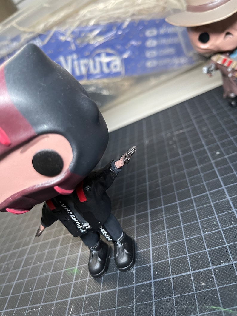 May include: A black and red vinyl figure wearing a black hooded jacket and black trousers. The figure has a black and white graphic on the jacket that says "Unbreakable". The figure is wearing black shoes.