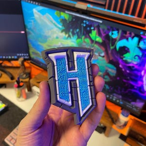 Puede incluir: Un emblema gris, blanco y azul con forma de letra "H". La letra está rellena con un patrón azul detallado y delineada en blanco. El emblema tiene un borde gris.