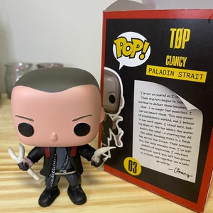 May include: A Funko Pop! vinyl figure of Clancy, a character from the TV show "Top: Paladin Strait". The figure is wearing a black jacket with a red collar and a white shirt. He is holding two white bone-like objects in his hands. The figure is standing on a wooden surface. The background is a box with the Funko Pop! logo and the character's name and show title.