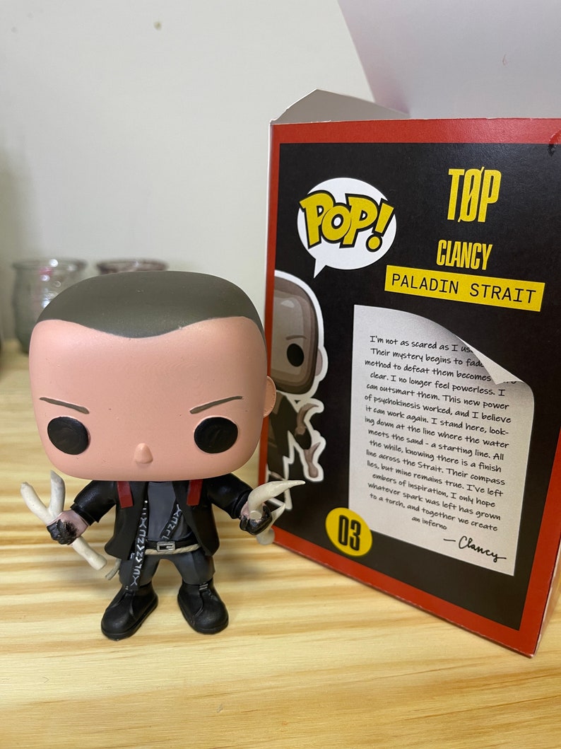 May include: A Funko Pop! vinyl figure of Clancy, a character from the TV show "Top: Paladin Strait". The figure is wearing a black jacket with a red collar and a white shirt. He is holding two white bone-like objects in his hands. The figure is standing on a wooden surface. The background is a box with the Funko Pop! logo and the character's name and show title.