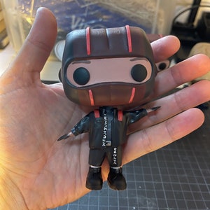 May include: A brown and red pop vinyl figure with a black mask and black clothing. The figure is wearing a black jacket with white text that reads "TRUJAZUNコ" and "CANUX".