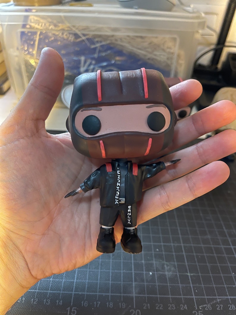 May include: A brown and red pop vinyl figure with a black mask and black clothing. The figure is wearing a black jacket with white text that reads "TRUJAZUNコ" and "CANUX".